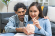 © KMPZZZ - Happy smile asian young couple love hand putting coin in piggy bank for saving money to buy real estate for new home. Business finance, deposit with banking for financial planning future together.