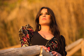 Wall Mural - Spanish woman, brunette and beautiful, dancing flamenco with typical flamenco dancing costume leaning on a wooden railing and looking at the sea at the horizon at sunset. The woman is sad.