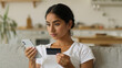 © Yuliia - Online internet payment serious Indian woman Arabian female ethnic girl at home Hindu lady looking at mobile phone using smartphone holding gadget buying with credit card bank app shopping loan credit
