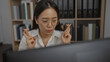 © Krakenimages.com - Young chinese woman with glasses crossing fingers in an office setting, surrounded by books and file cabinets, focused on a computer screen.
