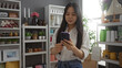 © Krakenimages.com - Young chinese woman browsing phone in a beautifully decorated home decor store with shelves of various items including plants, jars, and ornaments.