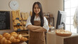 © Krakenimages.com - Young woman with long dark hair in a brown apron standing confidently with folded arms in a bakery shop filled with various freshly baked bread displayed around her