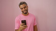 © Krakenimages.com - Young arab man with a beard, wearing a pink shirt, smiling while looking at his phone against an isolated pink background.