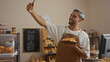 © Krakenimages.com - Handsome young man taking a selfie while holding croissants in a cozy indoor bakery shop with various pastries on display.