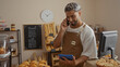 © Krakenimages.com - Young man with beard in bakery shop talking on phone while holding a tablet, dressed in apron, surrounded by pastries and bread, with a clock on the wall behind him