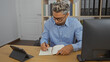 © Krakenimages.com - Young arab man working in his office writing in a notebook with a computer and tablet on his desk