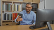 © Krakenimages.com - Young arab man with beard and glasses smiling while looking at his phone in a modern office setting with bookshelves in the background