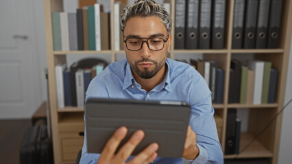 Young, arabian, bearded, man reading tablet in indoor office setting with bookshelves in the background