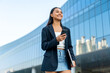 © StockPhotoRepublic - Young Asian Gen Z Businesswoman Using Phone While Carrying Laptop Walking in Modern Office District Outdoors