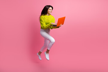 Wall Mural - Smiling young woman in a stylish outfit working on a laptop while happily jumping against a vibrant pink background