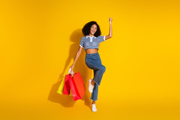 Young woman holding shopping bags joyfully against a vibrant yellow background, wearing casual stylish attire and expressing happiness