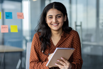 Wall Mural - Webcam view, woman looking into camera holding notepad for notes. Business meeting, online conference, employee happily writing down data while sitting inside office at table.