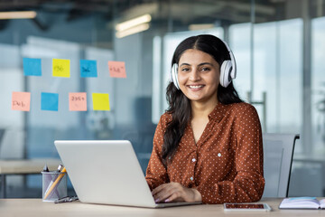 young woman working inside office wearing headphones, programmer smiling and looking at camera, empl