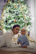 © saulich84 - A mother in a white sweater sits with her daughter for 1 year on the floor by the Christmas tree in an apartment on New Year's Eve