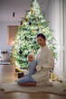 © saulich84 - A mother in a white sweater sits with her daughter for 1 year on the floor by the Christmas tree in an apartment on New Year's Eve