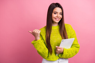 Wall Mural - Happy young woman wearing a lime green sweater holding a tablet and pointing with her thumb against a pink background