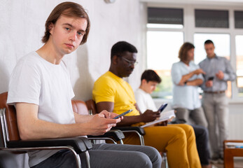 Wall Mural - Young male patient sitting on the bench in waiting room of clinic together with other guys