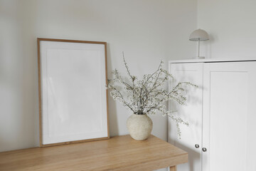 Wall Mural - Spring Scandinavian interior still life. Blank thin wooden picture frame mockup. White blossoming cherry plum tree branches in ceramic vase on bamboo table. White cabinets with modern portable lamp.