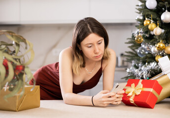 Wall Mural - Frustrated young woman at home against the background of a Christmas tree with presents, is typing a message to someone on .her mobile phone