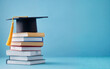 © zeenika - Graduation cap on stack of books against blue background symbolizing academic success and higher education achievement