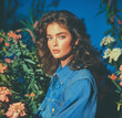 © Muhammad - A portrait of an attractive woman with wavy brunette hair, wearing blue denim shirt and posing in front of plants on the right side, with a royal blue background. She has soft glam makeup