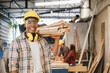 © sorapop - Smiling construction worker in a yellow hardhat and plaid shirt holding wood planks on his shoulder. Ideal for carpentry, woodworking, and craftsmanship in furniture workshop, National Carpenters Day