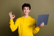 © deagreez - Young man in yellow sweatshirt holding laptop, showing OK gesture, standing in front of khaki background
