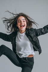 Wall Mural - Photo of a laughing young woman kicking her legs in the air, wearing a white t-shirt and a black jacket, on a gray background. The composition is simple, with a feminine and natural feel