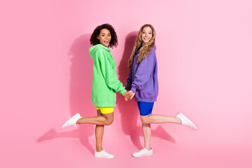 Canvas Print - Two cheerful women standing on a pink background in bright casual outfits, holding hands and showing their fun and playful friendship