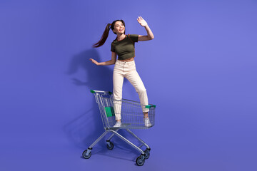 Wall Mural - Young woman posing cheerfully with a shopping cart on violet backdrop, emphasizing youthful fun and creativity