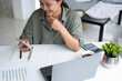 © MIND AND I - Businesswoman sitting at desk on couch in workplace or at home working on laptop and analyzing data on charts and graphs and writing on papers to make business plan and strategies for company