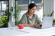 © MIND AND I - Businesswoman sitting at desk on couch in workplace or at home working on laptop and analyzing data on charts and graphs and writing on papers to make business plan and strategies for company