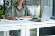 © MIND AND I - Businesswoman sitting at desk on couch in workplace or at home working on laptop and analyzing data on charts and graphs and writing on papers to make business plan and strategies for company