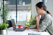 © MIND AND I - Businesswoman sitting at desk on couch in workplace or at home working on laptop and analyzing data on charts and graphs and writing on papers to make business plan and strategies for company