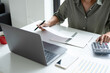 © MIND AND I - Businesswoman sitting at desk on couch in workplace or at home working on laptop and analyzing data on charts and graphs and writing on papers to make business plan and strategies for company