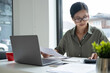 © MIND AND I - Businesswoman sitting at desk on couch in workplace or at home working on laptop and analyzing data on charts and graphs and writing on papers to make business plan and strategies for company