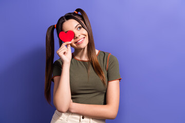 Wall Mural - Young woman with playful hairstyle holding a red heart shape while smiling against a purple background