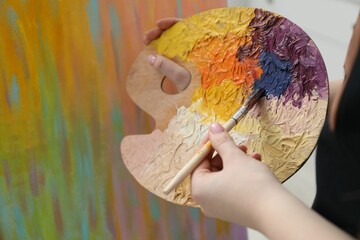 Wall Mural - Woman with paintbrush mixing paints on palette near picture, closeup