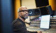 © Olena - African American man with glasses, sitting at the mixing console with computer monitor screen in a recording  music studio. Middle-aged Black male sound producer working in a modern Broadcast  studio.
