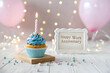 © PulseImages - Work anniversary cupcake with blue frosting, lit candle, and a greeting card on a festive table with soft pastel balloons in the background