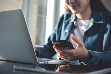 Wall Mural - Close up, young woman using mobile phone while working on laptop computer in office. Casual business woman, freelancer online working at home office. Student studing online, e-learning