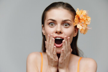 Wall Mural - Excited young woman with flower in hair, expressing surprise and joy in vibrant orange attire, against a soft gray background.