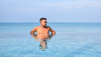 Wall Mural - A man enjoys a serene moment, soaking in the refreshing infinity pool overlooking the stunning blue sea of Krabi Thailand. The sun glistens on the water, creating a perfect getaway.