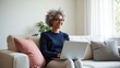 © saranya - cheerful older woman with curly gray hair and glasses, sitting on a cozy couch with a laptop.