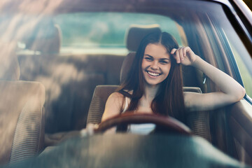 Wall Mural - Happy young woman driving a car, smiling confidently, showcasing freedom and adventure on a sunny day. Bright, cheerful mood with a blurred landscape background.