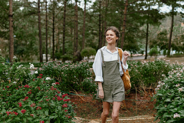 Wall Mural - Young woman enjoying a sunny day in a lush garden, wearing casual overalls and smiling while carrying a bag, surrounded by vibrant flowers and greenery