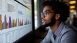© k - Un joven analista con gafas observa atentamente gráficos y datos en una pantalla grande. Su expresión refleja concentración en un entorno de trabajo moderno
