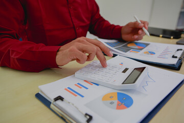 Wall Mural - An Asian businessman in a red shirt sits thoughtfully at his desk, and analyzing charts, deep in contemplation about maximizing profit and strategizing for future business growth.