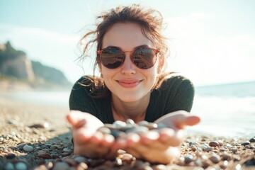 Wall Mural - an attractive woman in sunglasses, lying on the beach and picking up pebbles with her hands. She is smiling at the camera. 