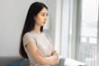 © fizkes - Young serious Asian woman stands with arms crossed in living room, gazing thoughtfully out window, deep in thought, reflecting, contemplating decision, moment of introspection, solitude, mindfulness